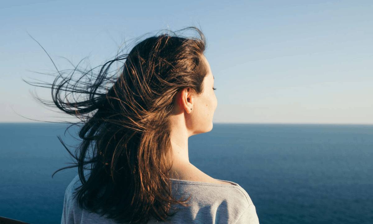 A woman looking out over the ocean, with her hair blowing in the wind beneath a clear blue sky.