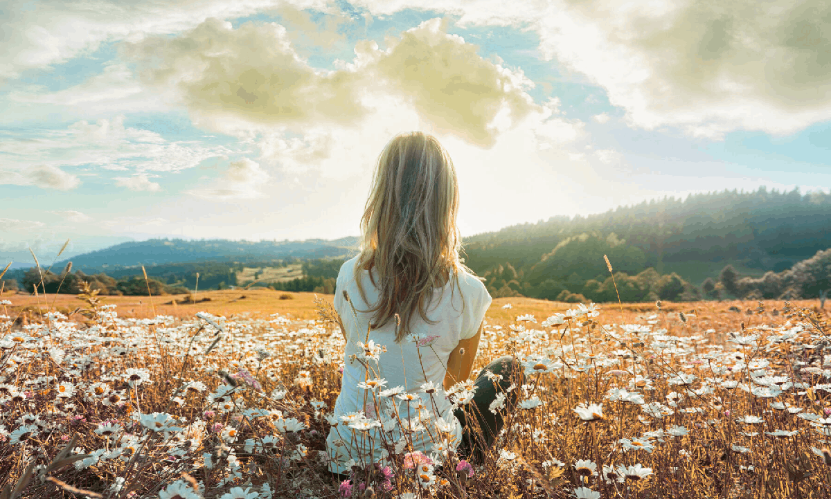 A woman sitting in a field of white flowers, looking out towards distant hills beneath a bright, cloudy sky.